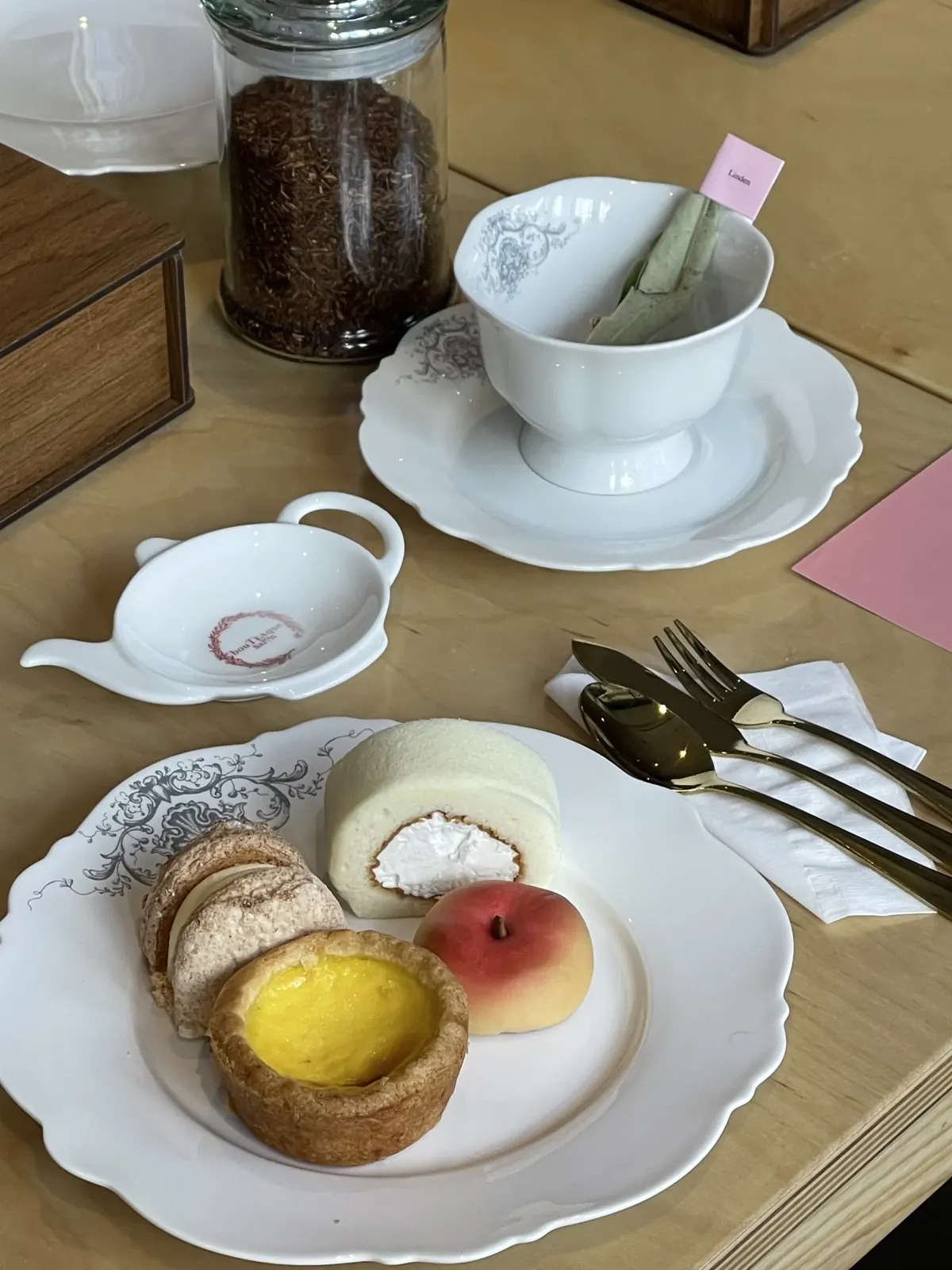 'Linden' 티백 태그가 꽂힌 컵, boutTEAque salon 로고가 새겨진 티포트 받침, 디저트 플레이트 클로즈업