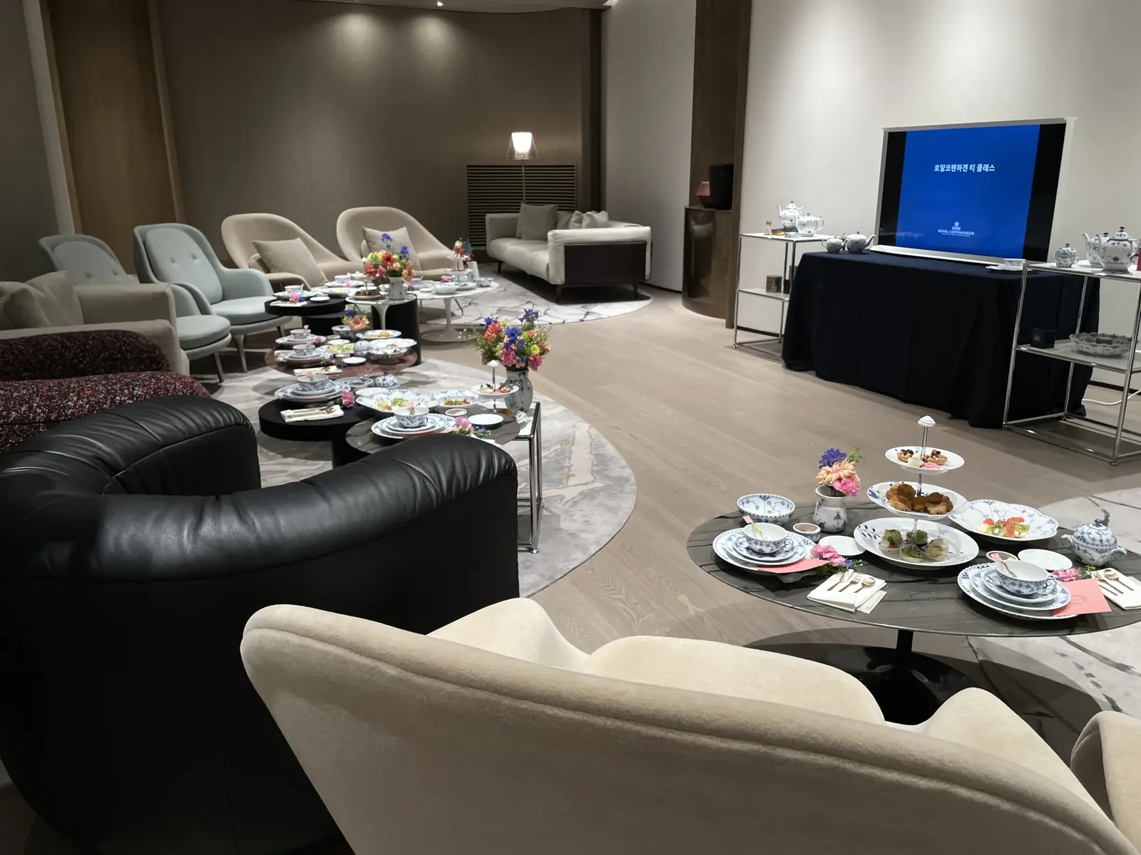 롯데백화점 동탄점 로얄코펜하겐 라운지 광각샷 — 다수의 좌석과 원형 테이블, TV 슬라이드