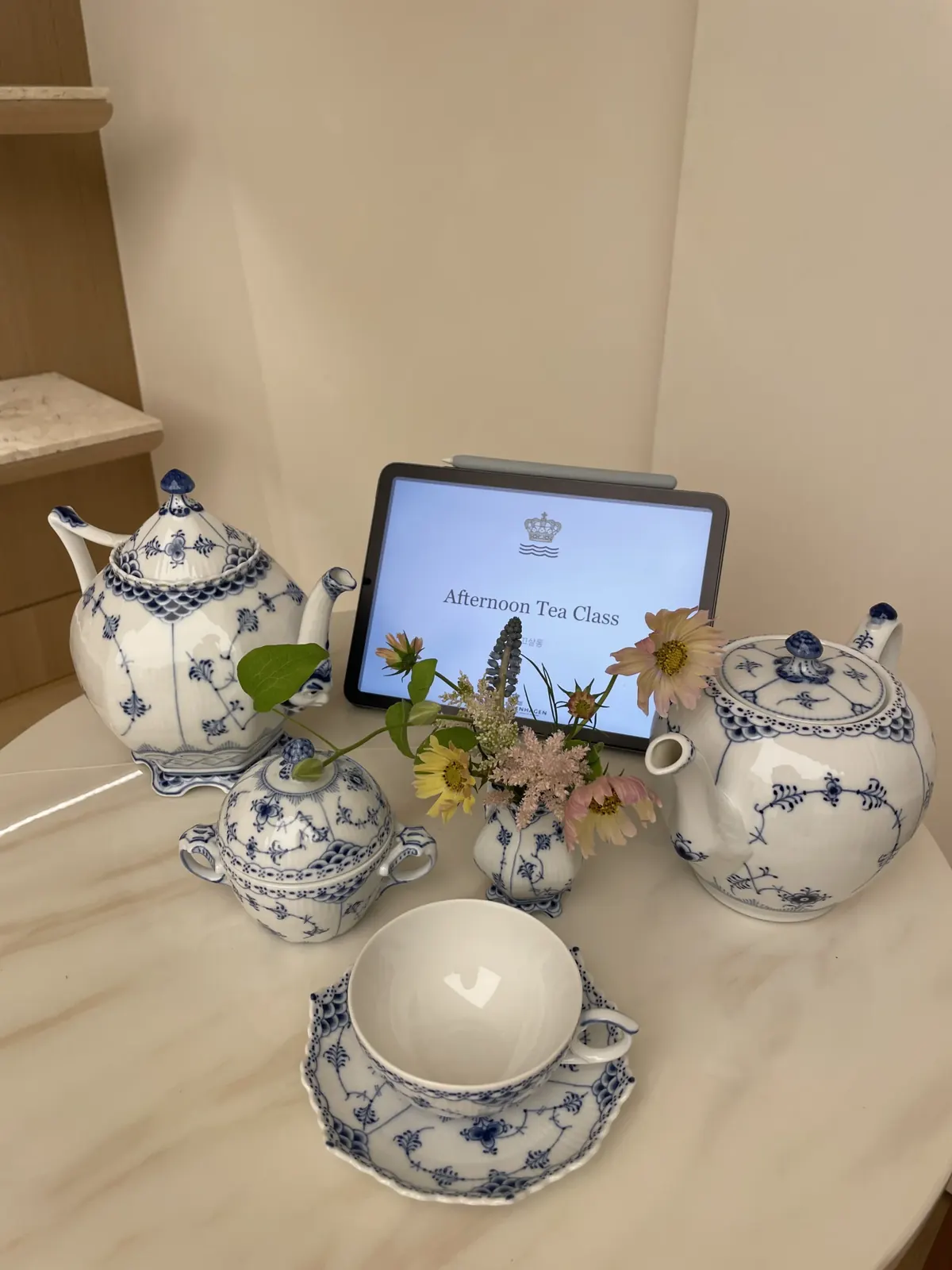 블루 플루티드 티팟과 슈가볼, 찻잔 세트 옆에 'Afternoon Tea Class' 문구가 뜬 태블릿이 놓인 테이블 세팅