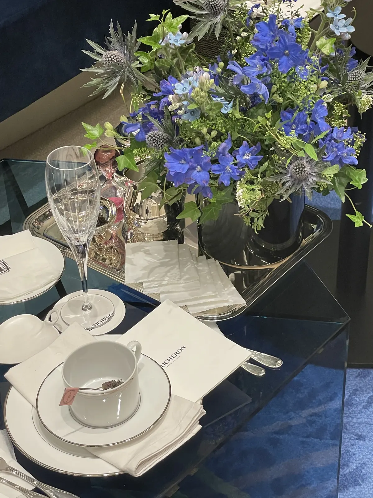 'BOUCHERON' 로고가 새겨진 냅킨과 백금테 찻잔, 은쟁반 위 샴페인 글라스, 블루 델피니움 센터피스