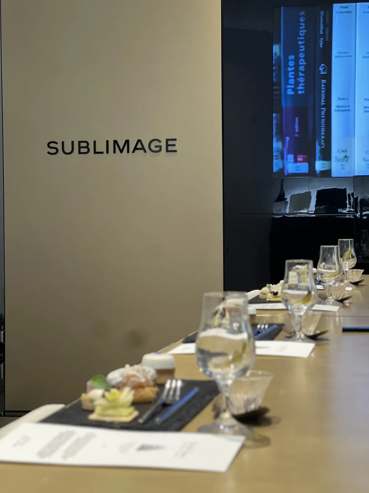 SUBLIMAGE 사이니지 벽과 책장이 보이는 스크린 앞, 여러 자리에 반복 세팅된 티페어링 테이블