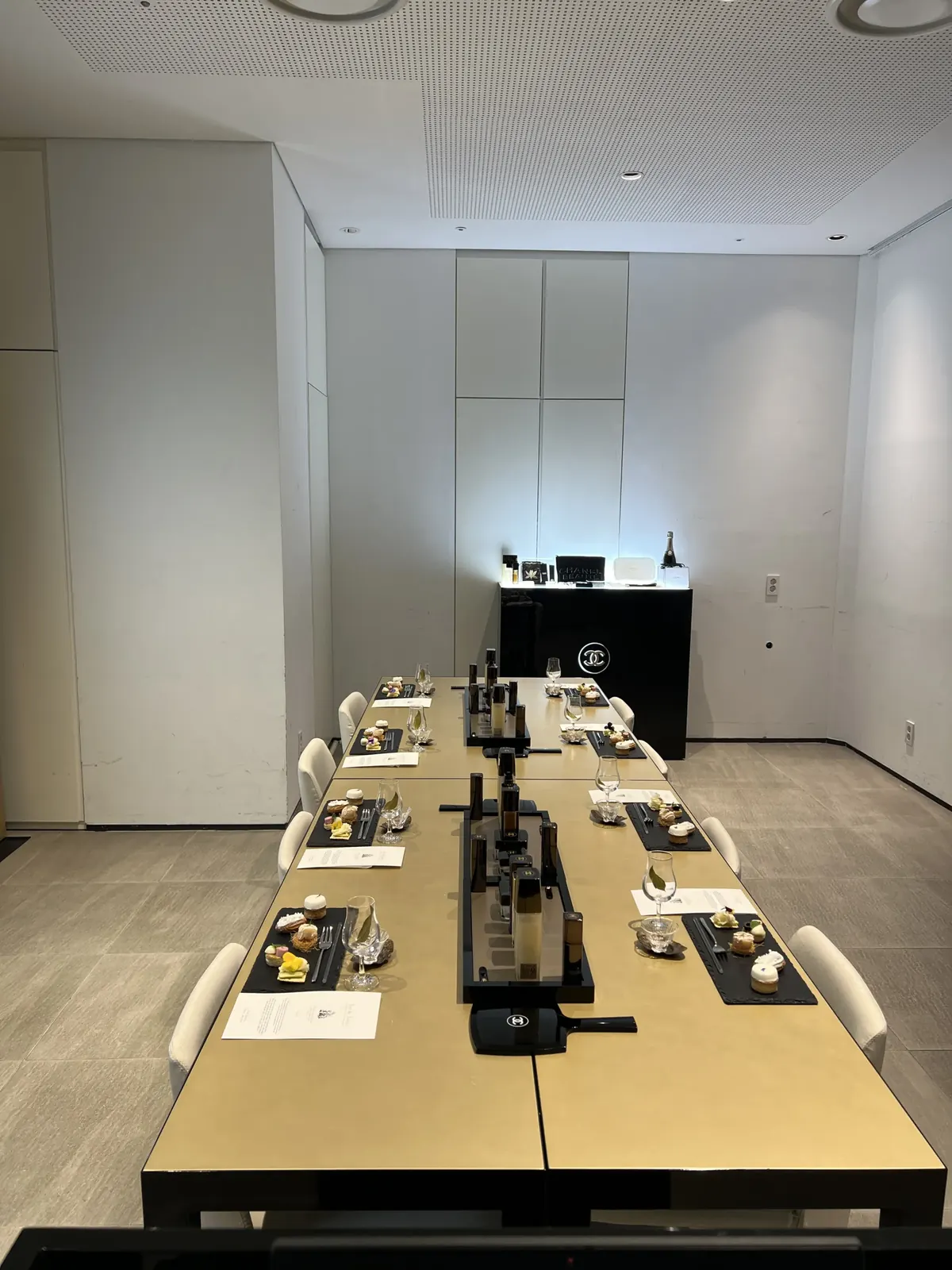 CHANEL CC 로고 조명 패널과 샴페인이 놓인 배경, 대칭형 긴 테이블에 반복 세팅된 시음 자리