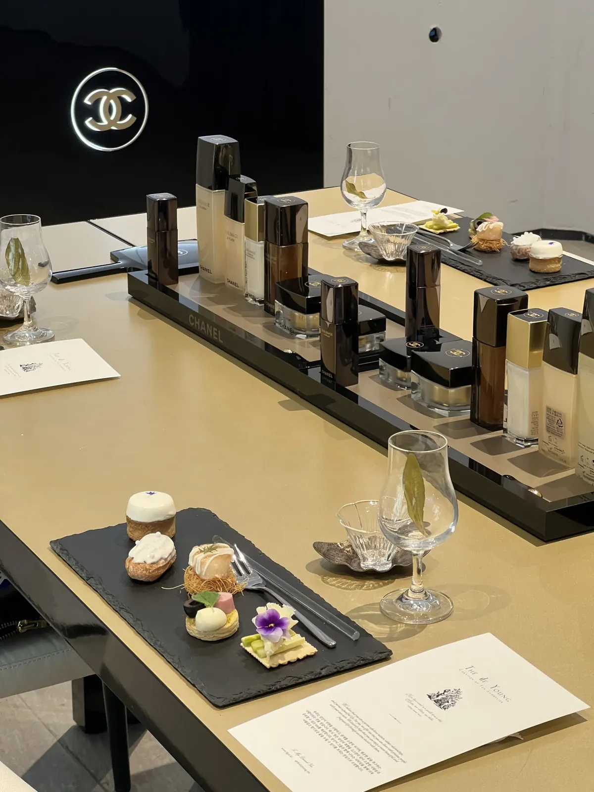 CHANEL CC 로고 조명과 브랜드 제품 라인업, THE de YOUNG 카드와 프티푸르가 함께 놓인 테이블