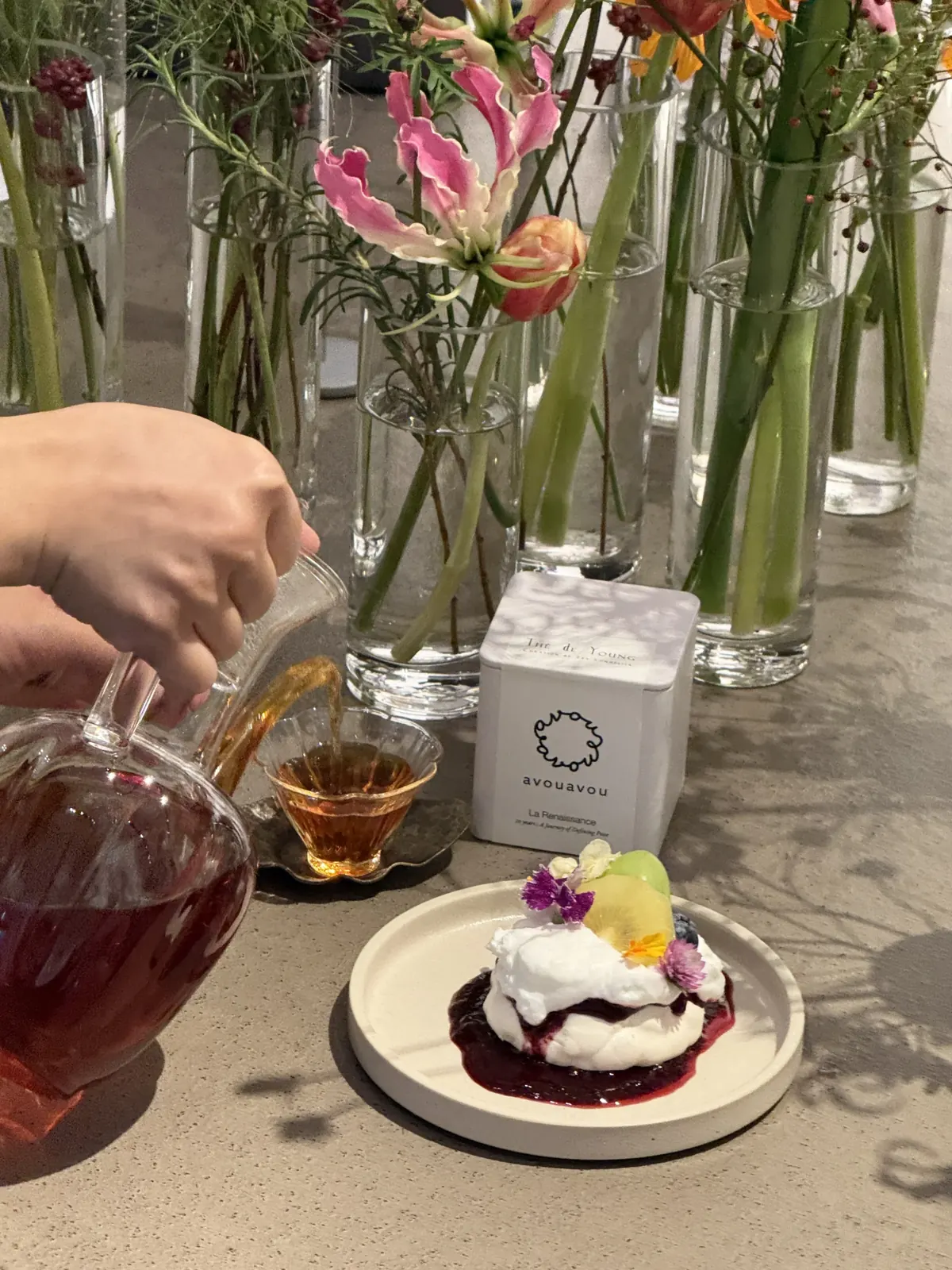 손으로 유리 카라프에서 잔으로 차를 따르는 동작, THE de YOUNG 박스와 디저트가 함께 놓인 장면